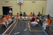 Photo d'entraînement d’initiation à la Capoeira de manière inclusive. La Photo montre des enfants qui s'assissent en rond dans un gymnase. Au milieu il y a une maitresse qui explique le tambourin.